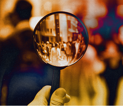 A hand holds a magnifying glass that focuses on a crowd of people